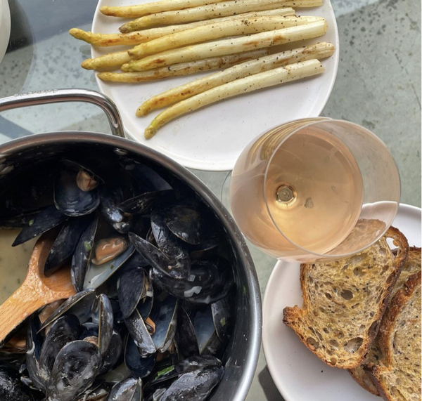 A large pot of black mussels beside a plate of white asparagus, toasted brown bread and a glass of rosé